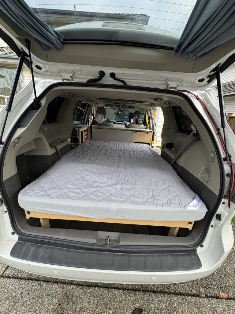 Tri-fold foam mattress with a grey mattress protector laid flat in bed mode on a plywood platform in a Toyota Sienna minivan camper