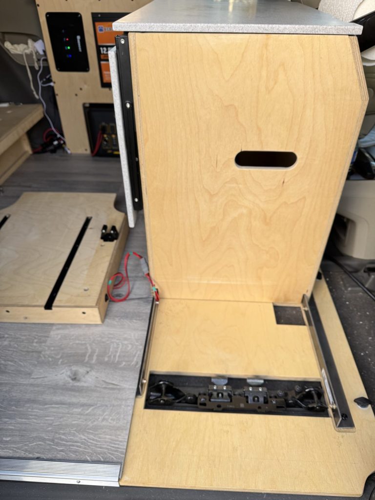 Plywood cabinet module mounted on the minivan camper floor next to the seat rail cutout and vinyl tiles