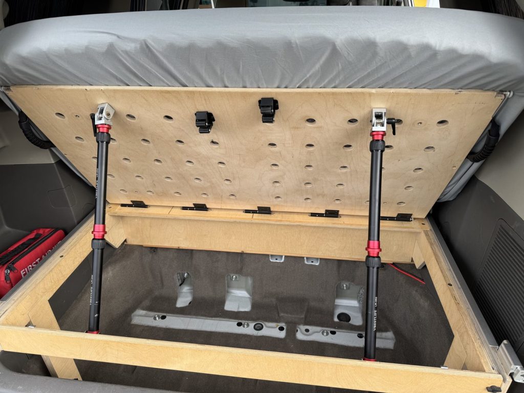 Head section of the minivan camper bed lifted into lounge mode, held by two black and red telescopic support legs above the open bed frame