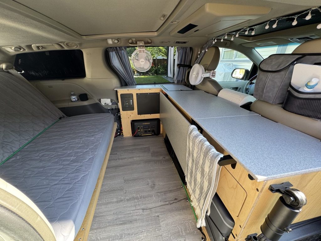 Camper bed folded into sofa mode with a grey quilted cover inside a Toyota Sienna minivan camper, next to the electrical panel and counter