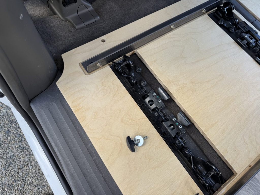Steel angle with rivet nuts on the plywood floor beside a seat rail cutout in a Toyota Sienna, with a knob bolt lying on the plywood