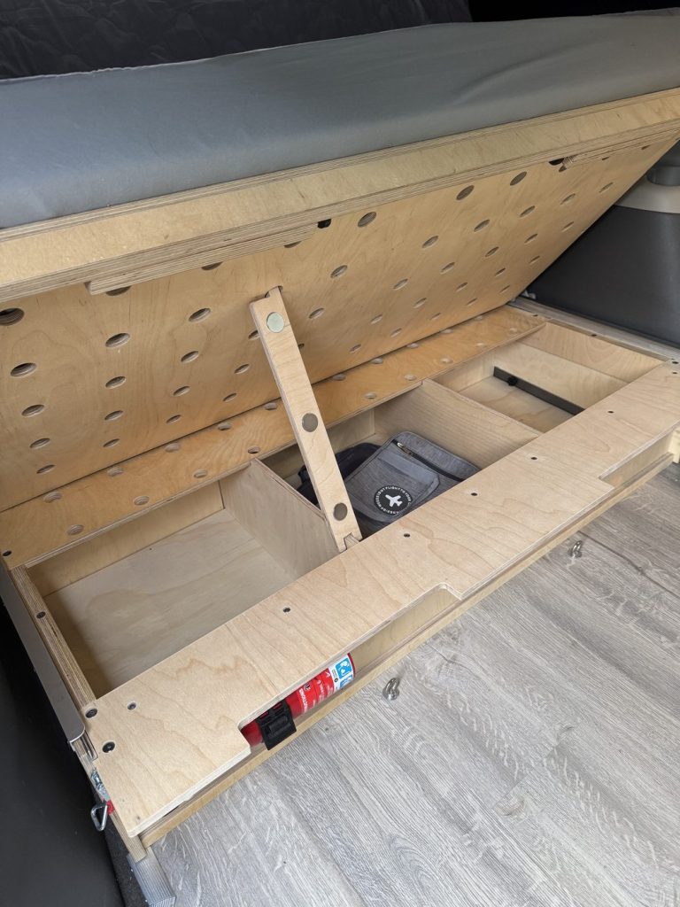 Under-bed storage box in a minivan camper with the seating section lifted and propped by a magnetic latch, showing divided compartments and a fire extinguisher