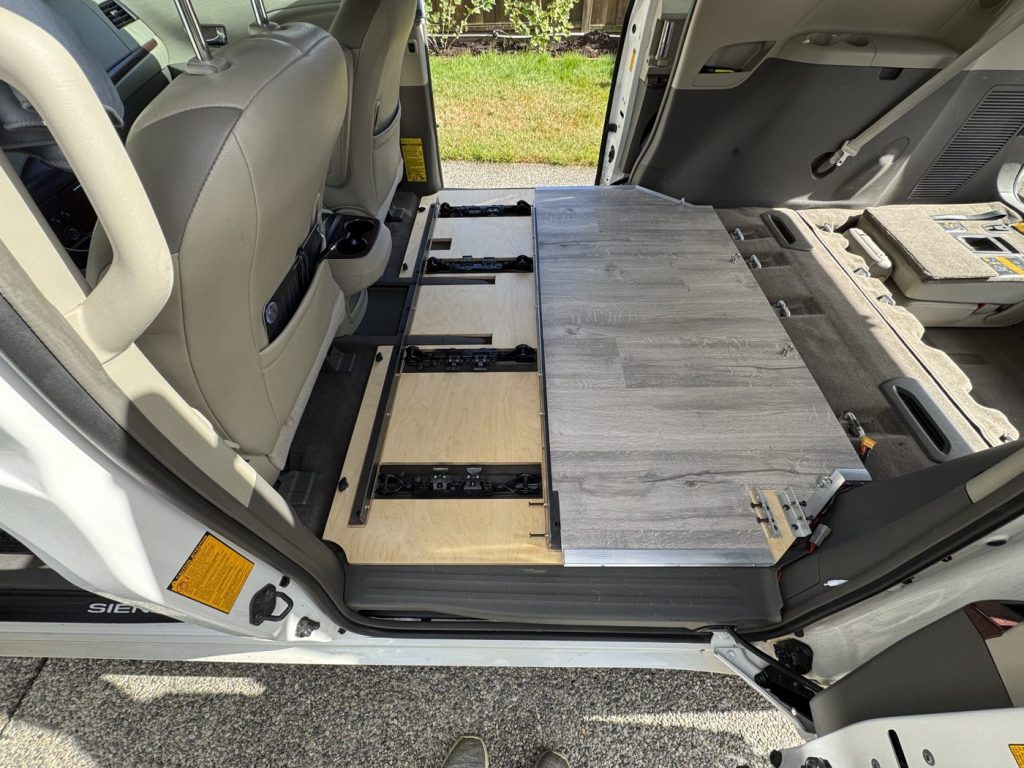 Grey vinyl tiles laid over half of the plywood subfloor in a Toyota Sienna minivan camper, with seat rail cutouts visible on the other half