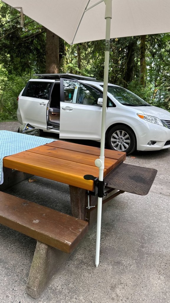 Cream umbrella attached with a black clamp to the edge of a wooden campground picnic table, with a white minivan camper parked behind it