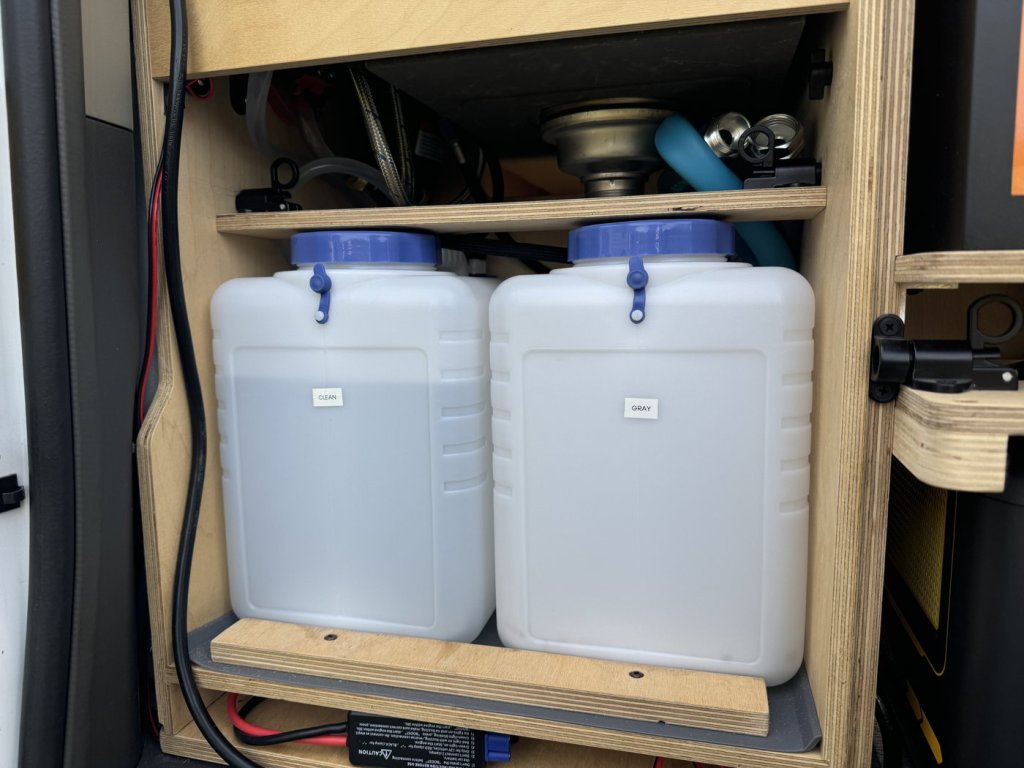 Labeled clean and gray 15-liter water tanks side by side in the minivan camper sink cabinet, held by a plywood barrier on a silicone mat