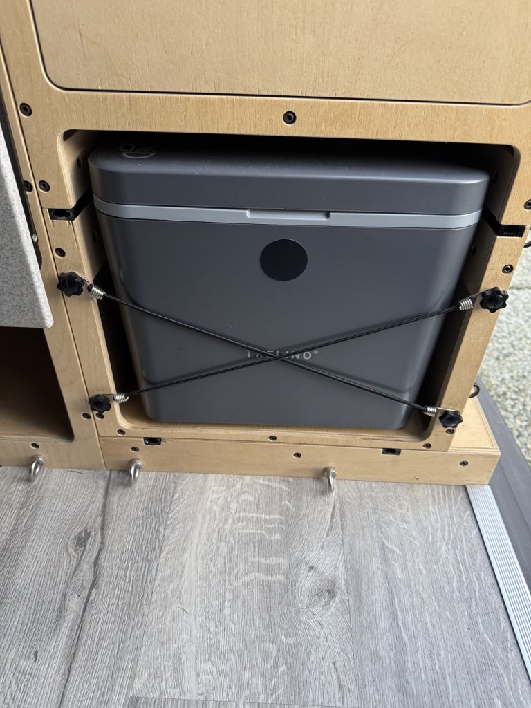 Separating composting toilet in the plywood storage unit of a minivan camper, held by two crisscrossed bungee cords