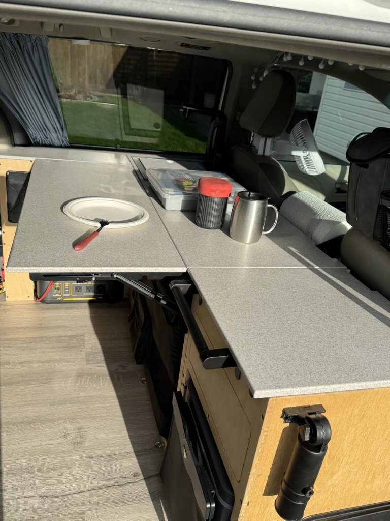 Unfolded Corian folding table extending the countertop of a minivan camper kitchen, with a plate, mug and paper towel holder