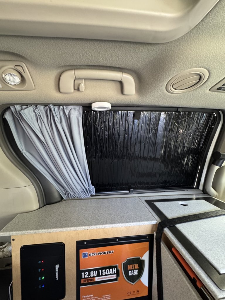 Grey curtain pulled to one side of a minivan camper window with a black WeatherTech blackout shade still in place, above the plywood kitchen module