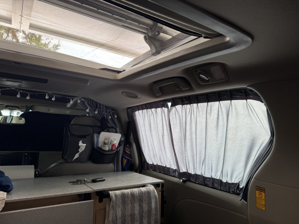Interior of a Toyota Sienna minivan camper looking toward the front, with curtains on the side windows, a divider curtain behind the front seats with string lights, and the sunroof open overhead
