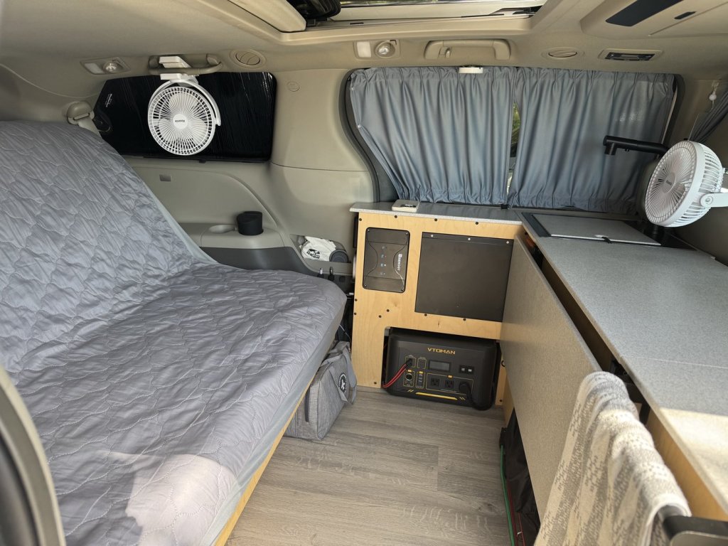 Inside a Toyota Sienna minivan camper with grey curtains drawn over the side window beside the kitchen module, a bed on the left and a black WeatherTech blackout shade on the opposite window