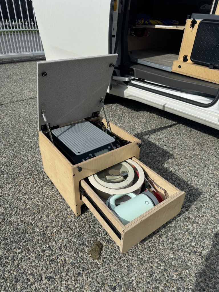 Detachable plywood cooking unit outside a Toyota Sienna minivan camper with the lid open, showing the induction cooktop and a drawer holding cookware and a kettle