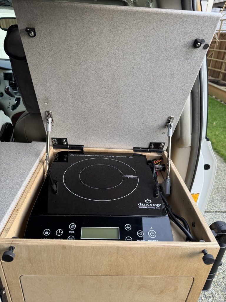 Duxtop induction cooktop mounted in a plywood cabinet module with a gas-strut lid raised, in a Toyota Sienna minivan camper
