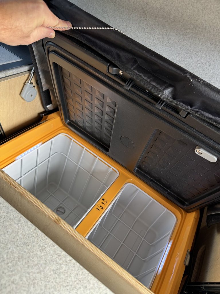 Hand pulling a ball chain to release the latch and open the lid of a dual-zone camper fridge inside a plywood cabinet