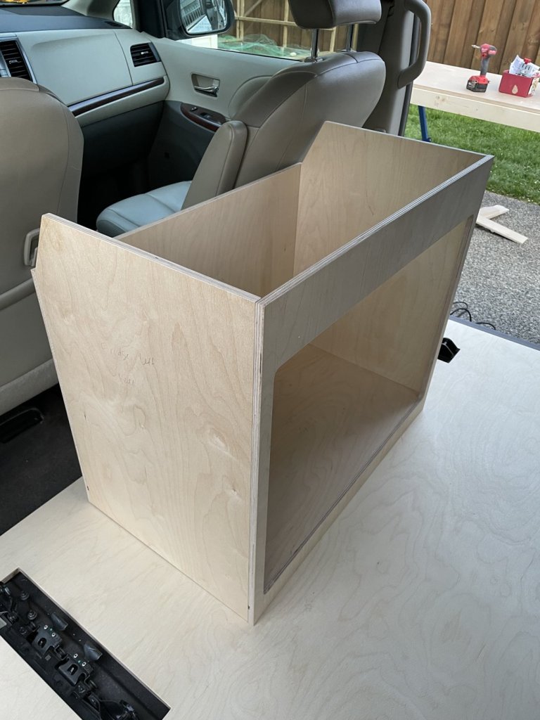 Bare birch plywood fridge cabinet with angled top and large front opening, placed next to the reclined front seat of a Toyota Sienna