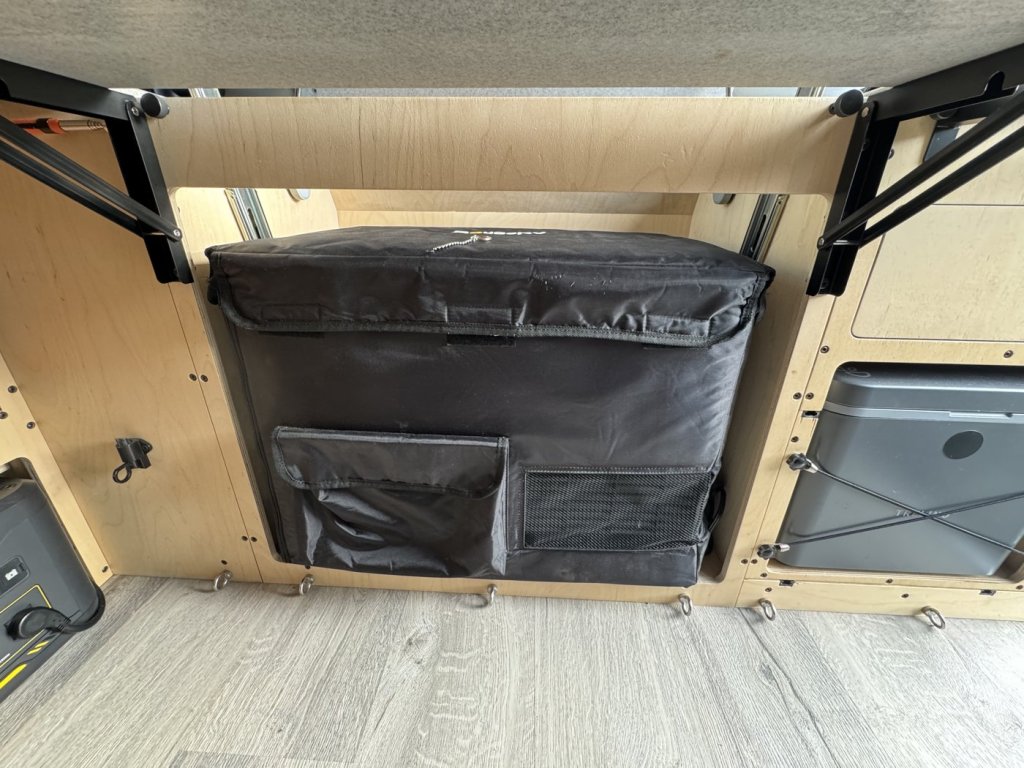 Black insulated cover on a 12V dual-zone camper fridge sitting in the plywood cabinet opening, with folding table arms above