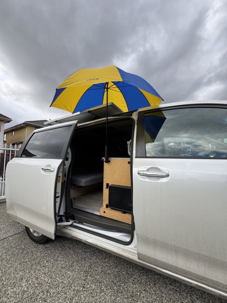 Blue and yellow IKEA umbrella held over the open sliding door of a Toyota Sienna minivan camper under a cloudy sky
