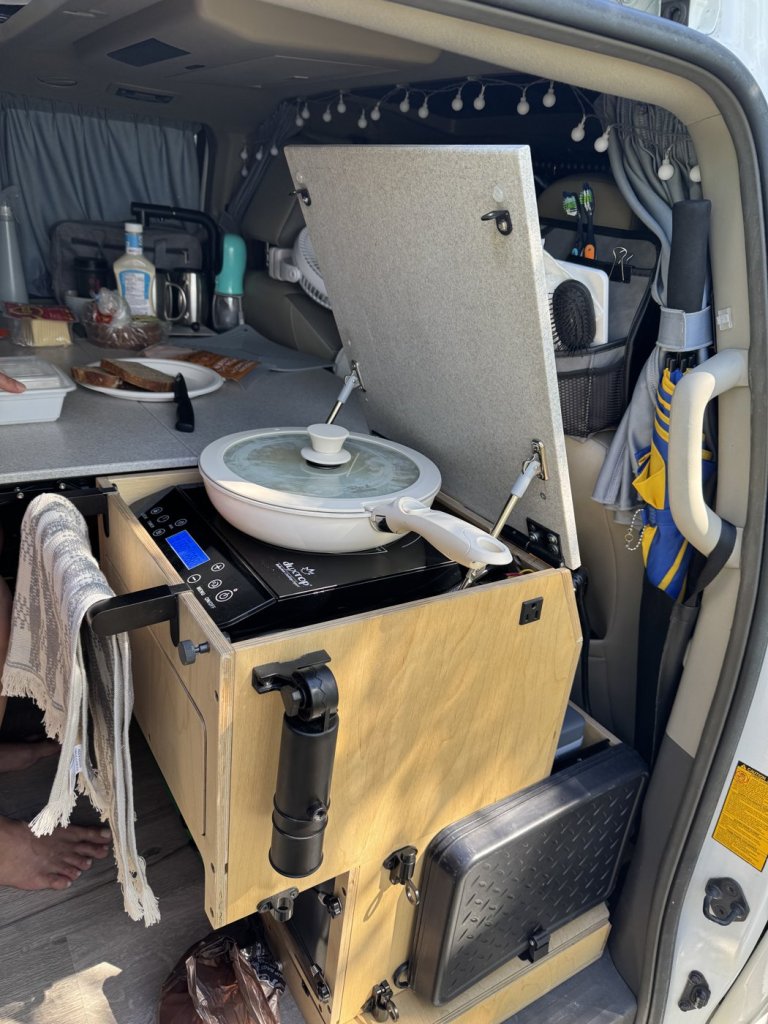 Frying pan with glass lid on a Duxtop induction cooktop inside a minivan camper, with lid open and a towel holder on the cabinet