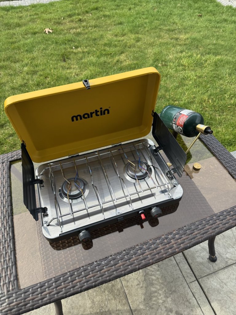 Yellow Martin portable dual-burner propane camping stove with wind guards open and a green propane cylinder attached, on an outdoor table