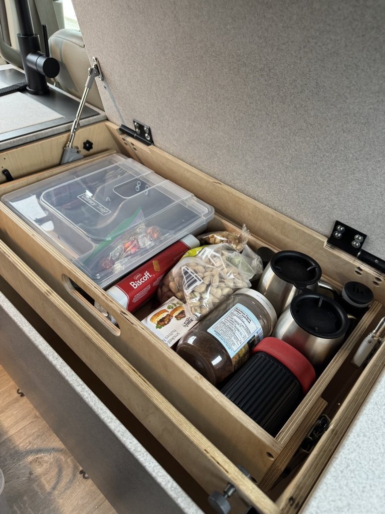 Open over-fridge storage drawer in a minivan camper holding snacks, a cutlery tray and travel mugs