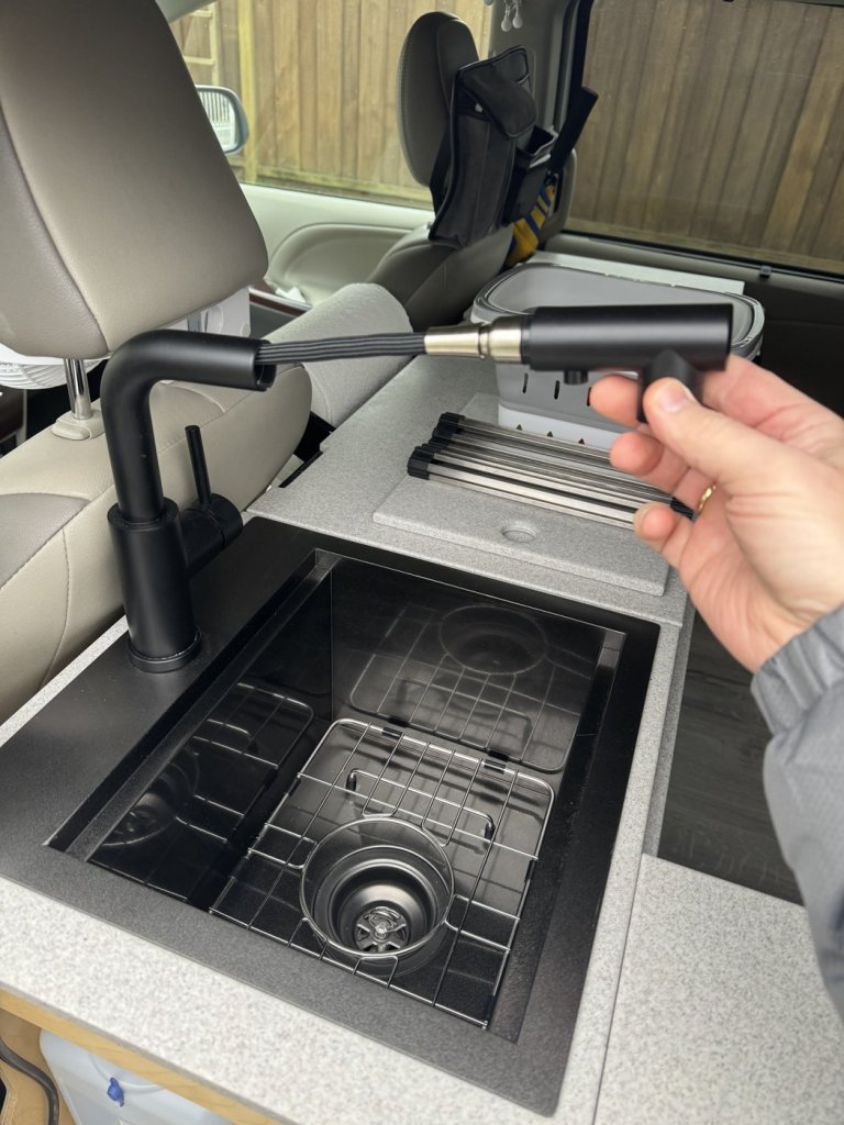Hand pulling out the extendable hose of a compact black faucet above the minivan camper bar sink with drain strainer and drying rack