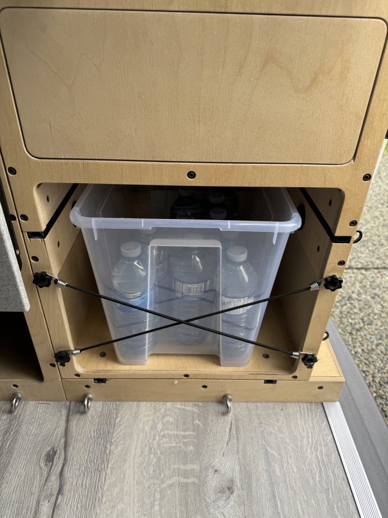 Clear storage bin of drinking water bottles held by crossed bungee cords in a rear side compartment of a minivan camper