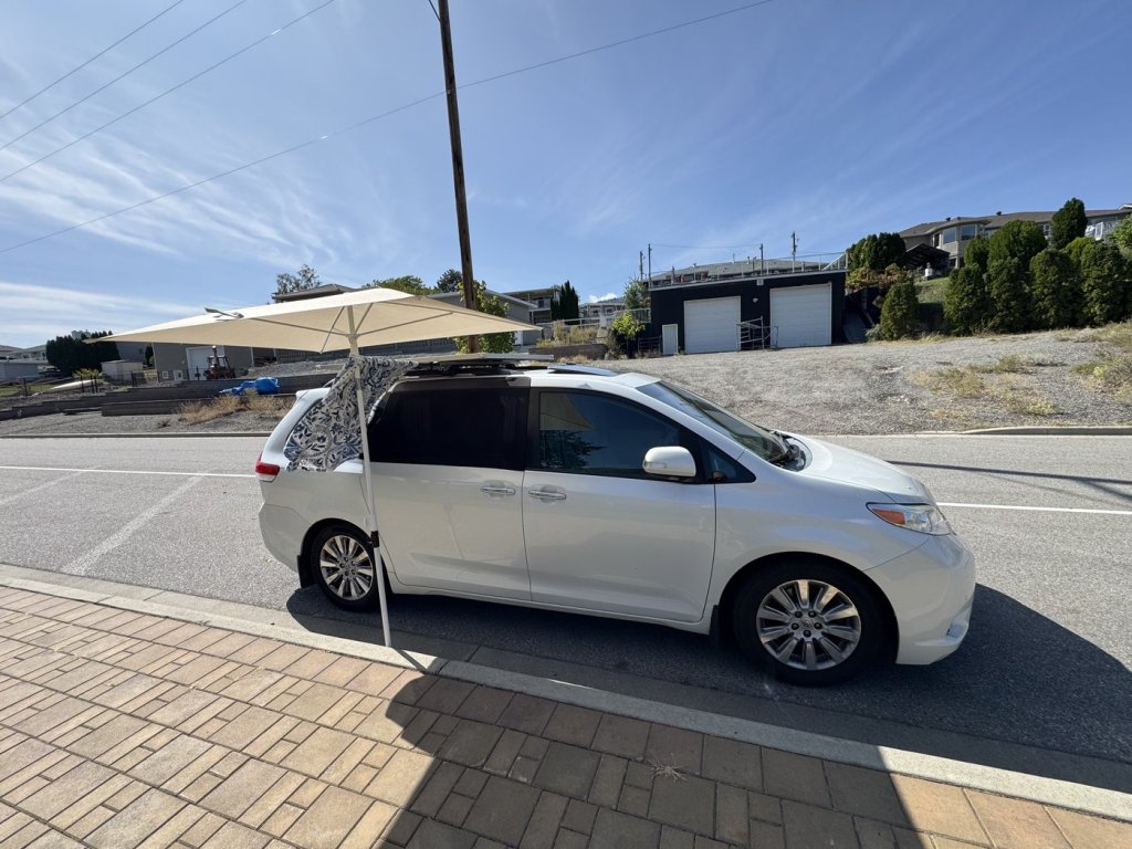Cream rectangular sun umbrella clamped to the roof crossbar under the solar panels of a white Toyota Sienna minivan camper parked on a street