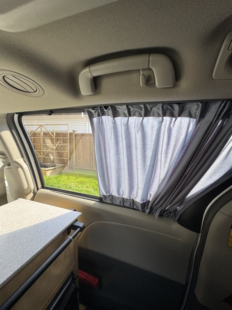 Grey curtain partly drawn across a Toyota Sienna minivan camper side window, with a garden fence and lawn visible through the open part of the glass