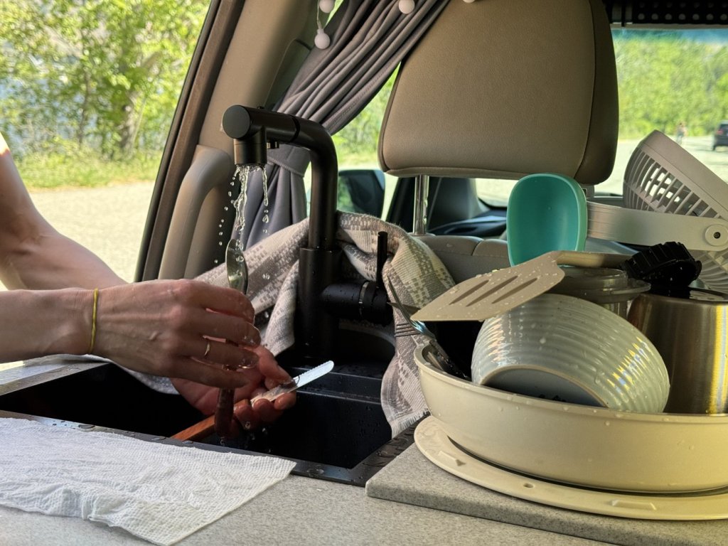 Washing cutlery under running water at the black faucet of the minivan camper sink, with dishes stacked on the countertop