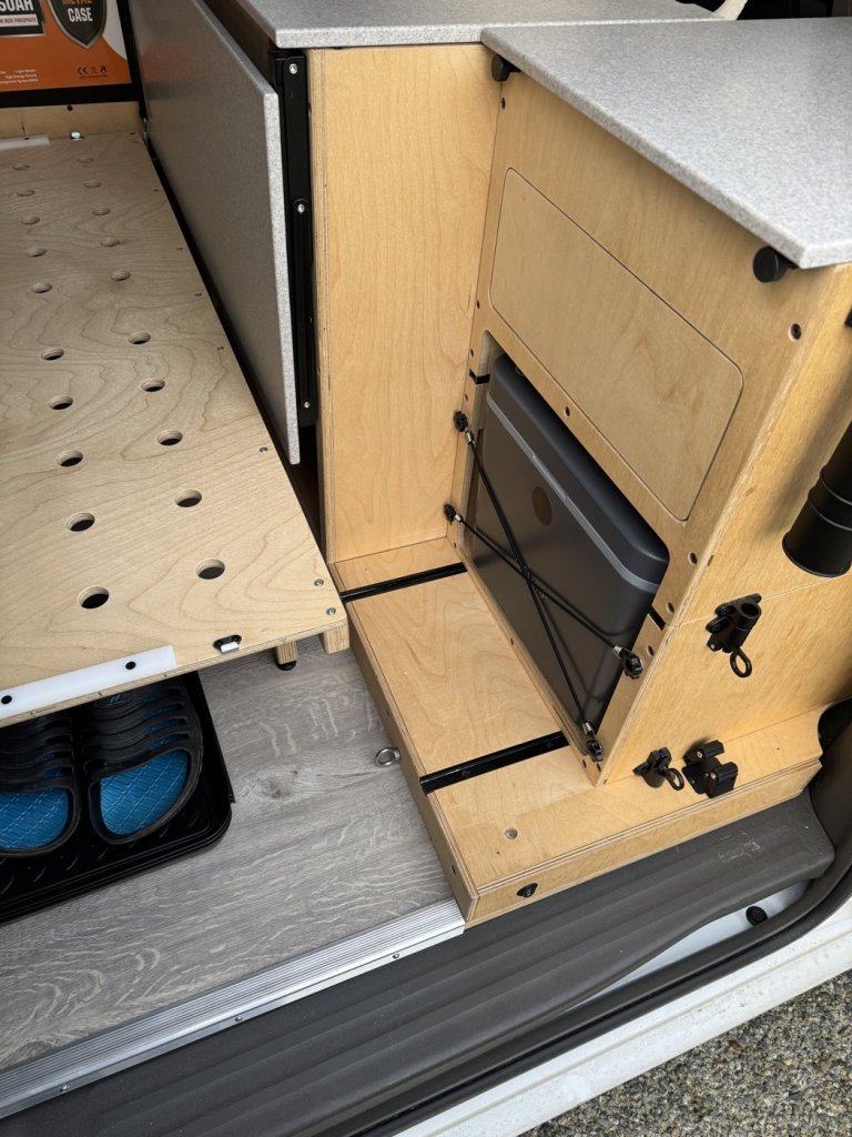 Base section of a minivan camper cabinet with black tracks and latches, with the storage unit slid toward the front of the van