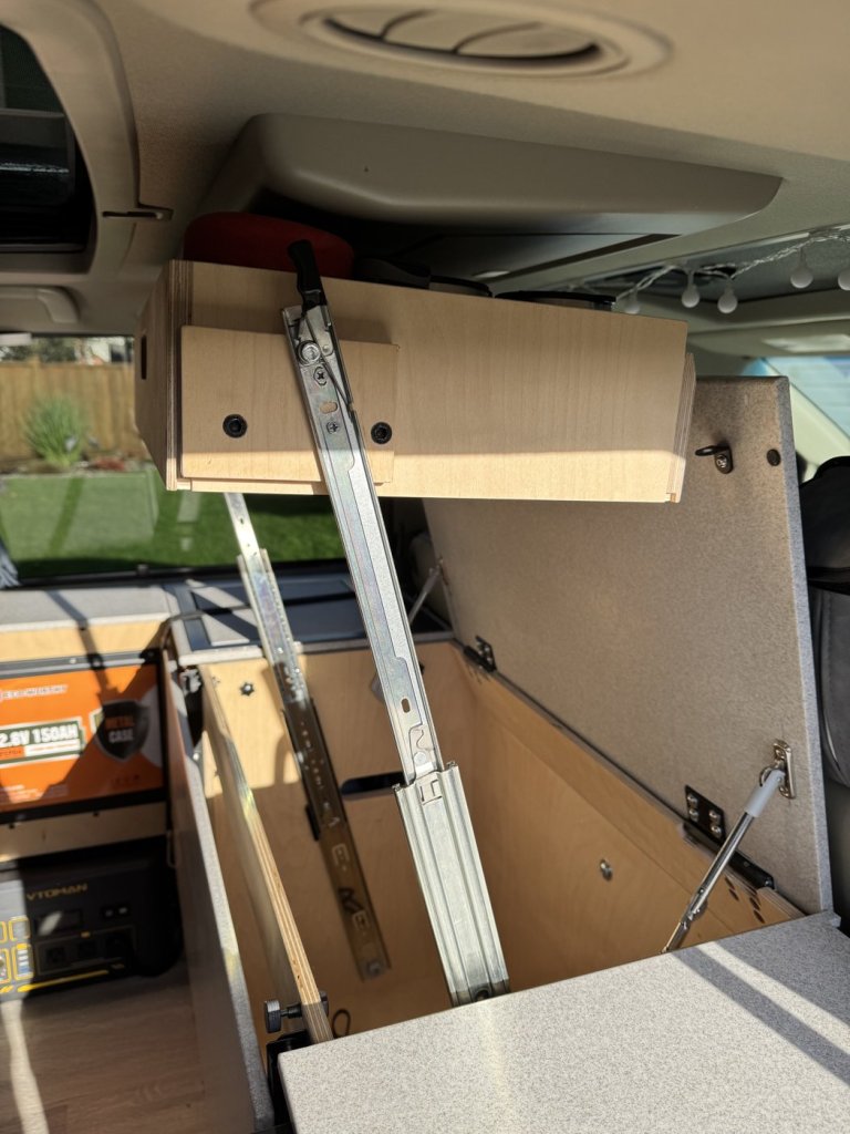 Plywood storage bin raised on metal rails above the fridge cabinet in a minivan camper, with the Corian lid held open
