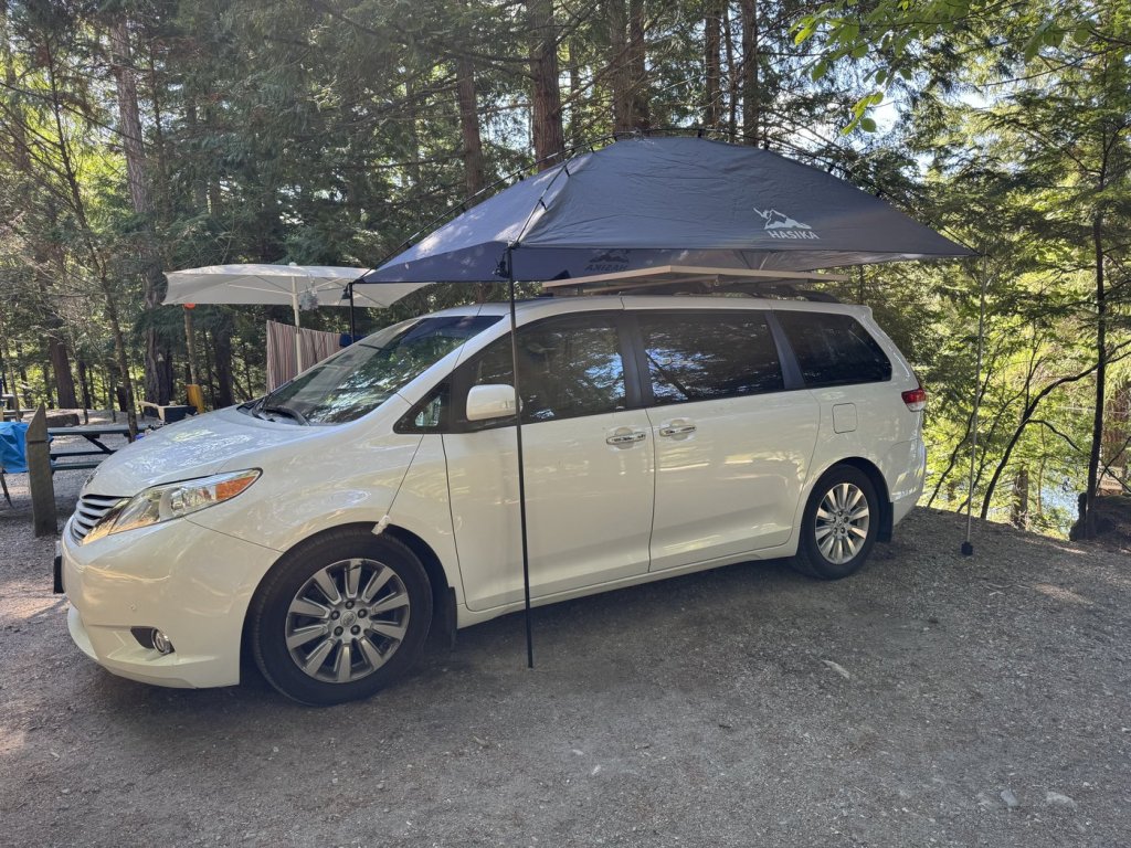Large dark teardrop awning set up over a white Toyota Sienna minivan camper at a forest campsite