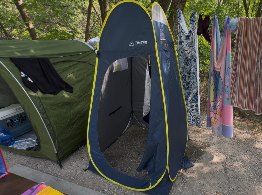 Navy blue TRATHM pop-up shower tent with yellow trim set up at a campsite, with towels drying on a line beside it