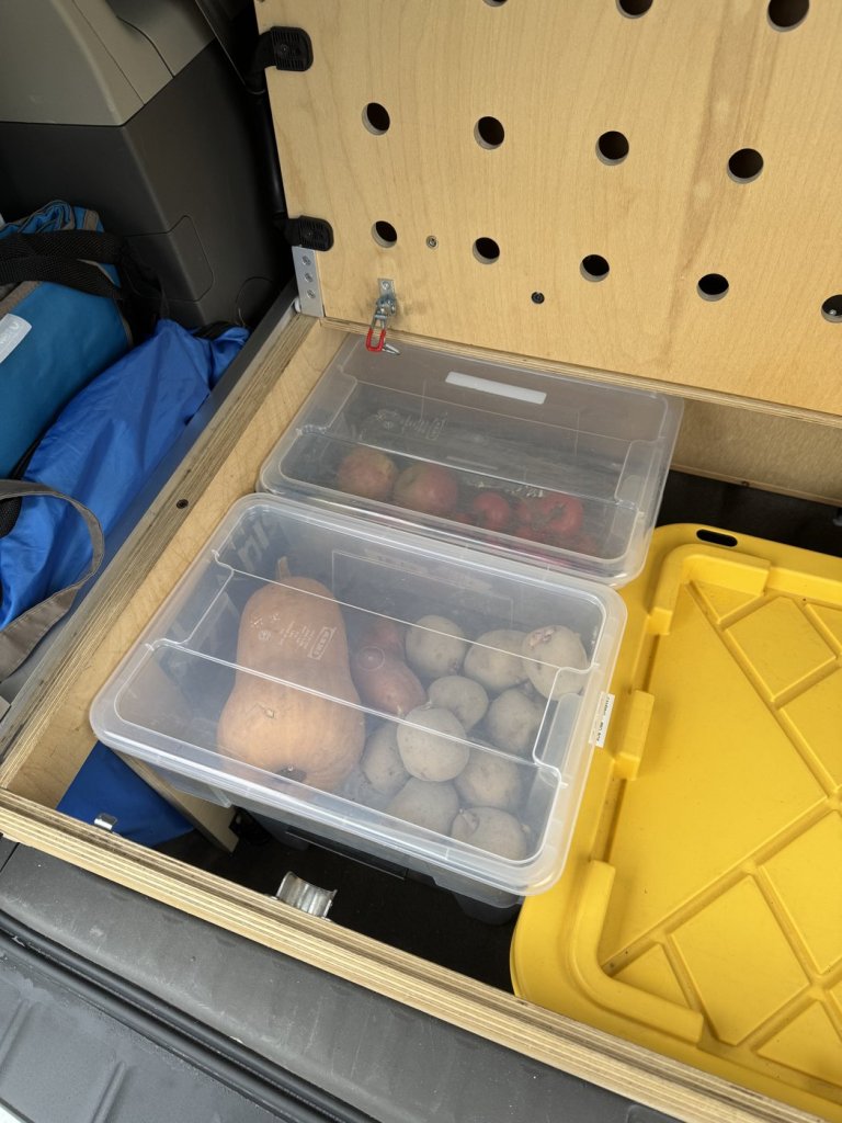 Clear boxes of vegetables and fruit next to a yellow storage tote in the trunk space under the bed of a minivan camper
