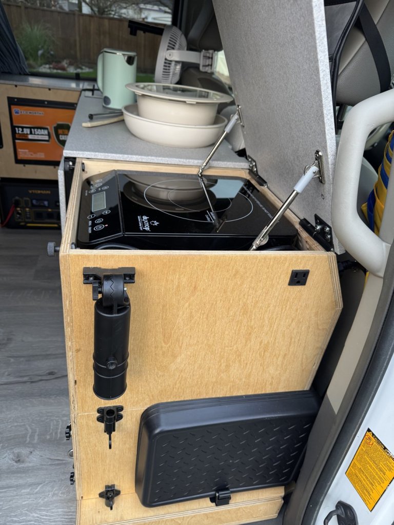 Side wall of a minivan camper cooking unit with a collapsible umbrella holder, a 120 V outlet and a black shoe tray holder