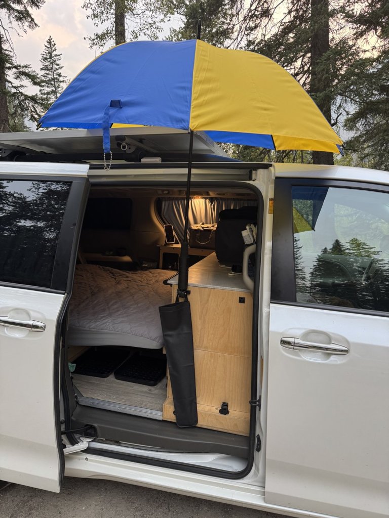 Open IKEA umbrella in a clamp holder on the cabinet beside the sliding door, with a black umbrella storage bag hanging below and the bed visible inside the minivan camper