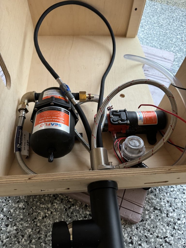 12V water pump, accumulator tank, clear inline filter and braided hoses laid out inside the plywood sink cabinet of a minivan camper