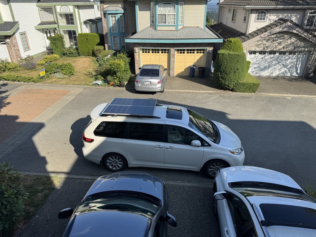 Toyota Sienna minivan parked on a residential street with two rigid solar panels bolted flat on the roof crossbars