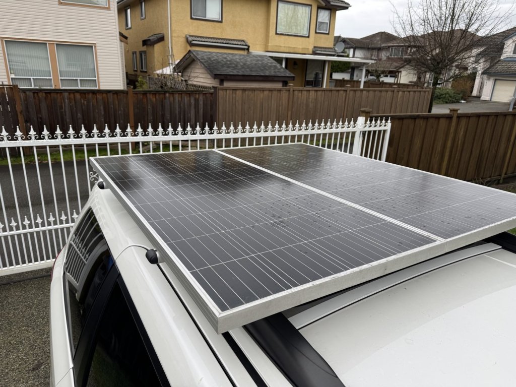 Close-up of two rigid solar panels mounted on crossbars above the roof of a white Toyota Sienna, raised over the moonroof